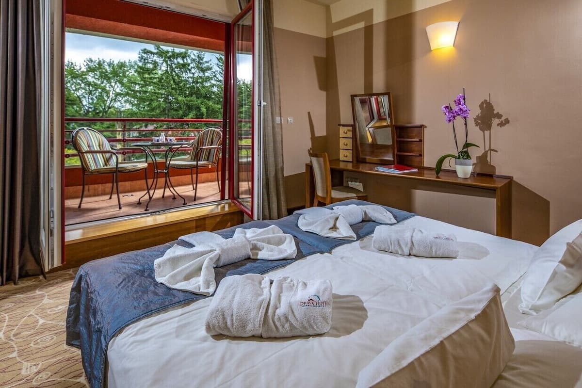 Ein Hotelzimmer mit Bett, Schreibtisch, Stuhl und einem Balkon mit Sitzgelegenheiten, mit Blick auf Bäume.