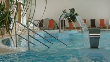 An indoor swimming pool with clear water, a clock on the wall, and orange lounge chairs around the pool.