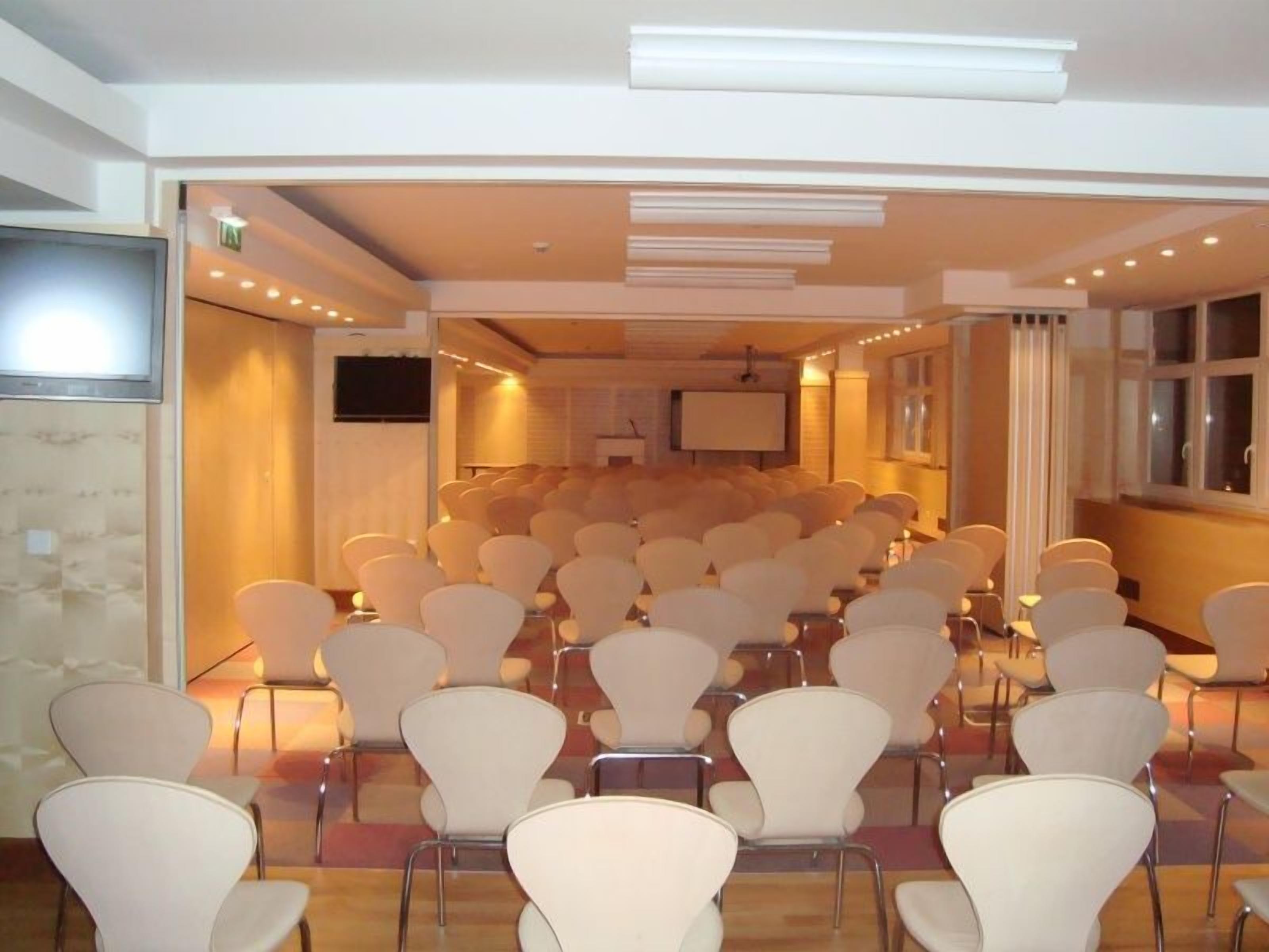Une salle de conférence avec des rangées de chaises blanches disposées face à une scène sur laquelle se trouve un écran.