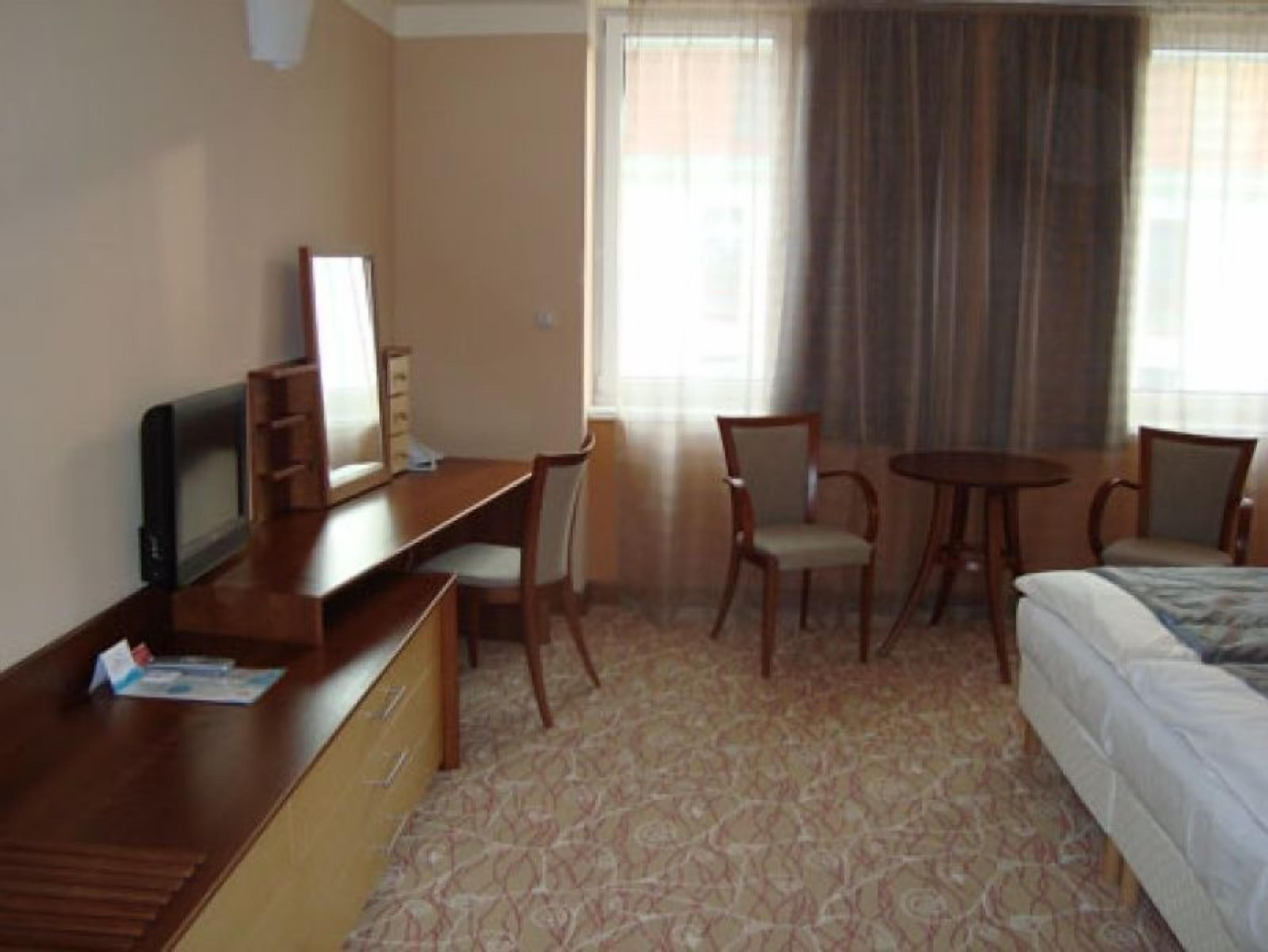 Een hotelkamer met een bed, een bureau met een computer, een stoel, een klein tafeltje en een raam met gordijnen.
