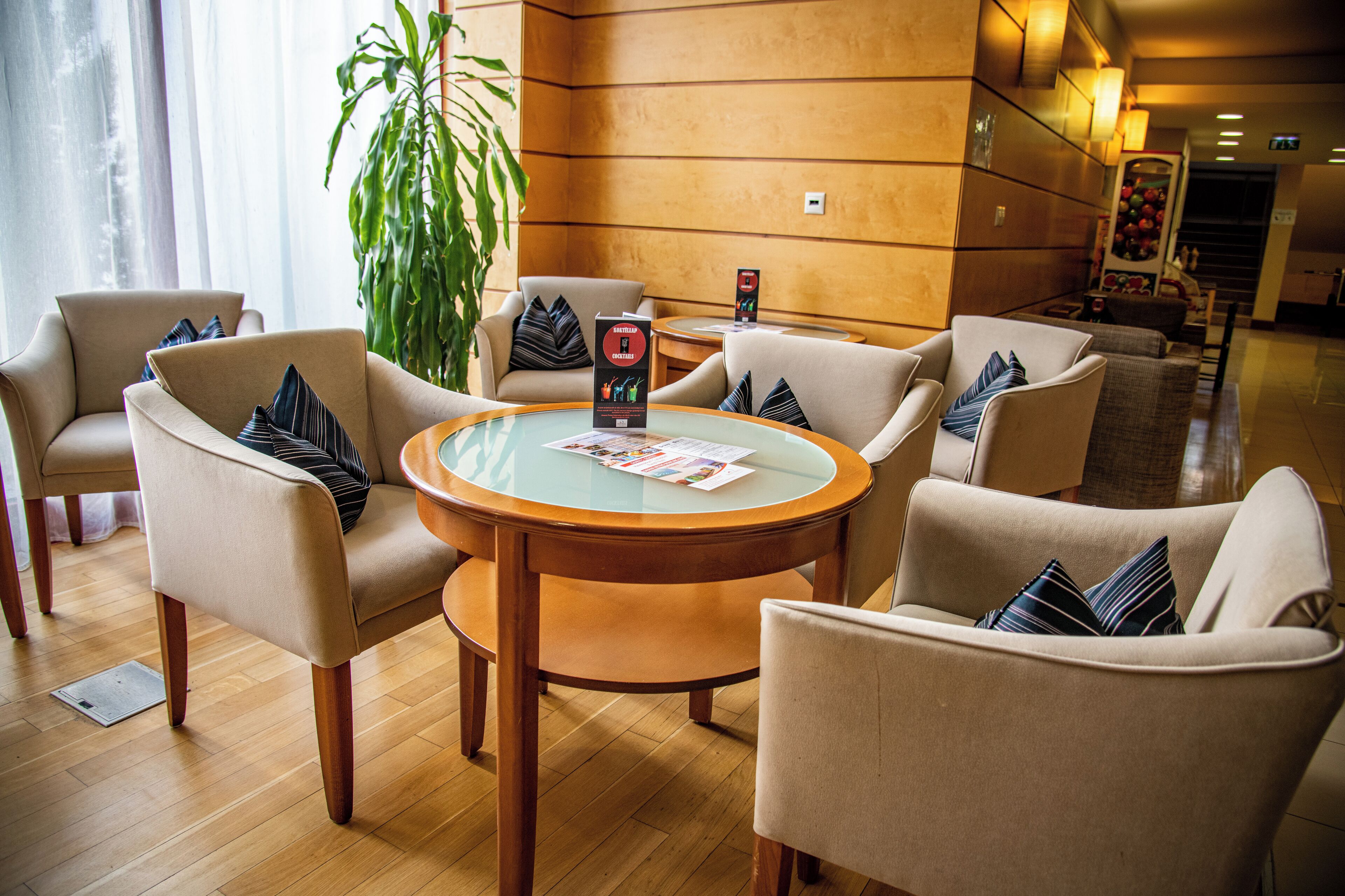 Een hal met een ronde houten tafel omringd door beige fauteuils, een grote kamerplant en een receptiebalie op de achtergrond.