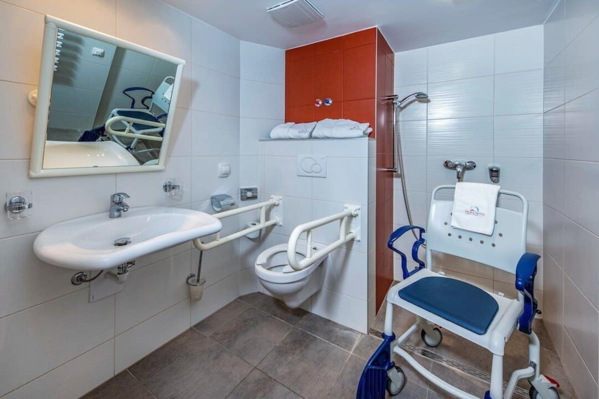 Ein Badezimmer mit weißem Waschbecken, Toilette inklusive Handrails, Duschbereich und einer rollstuhlgerechten Sitzbank.
