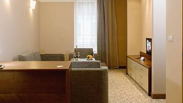 Une chambre d’hôtel avec un bureau en bois, un canapé gris, un petit pouf, une télévision et une commode.