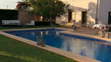 A pool area with lounge chairs and tables, a building with a tiled roof, and a tree in the background.