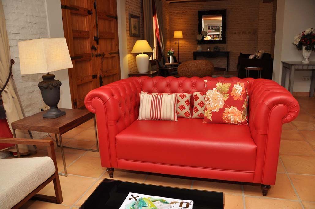 Un sofá de cuero rojo con cojines estampados en una habitación con pared de ladrillo, puerta de madera y una pequeña mesa con lámpara.