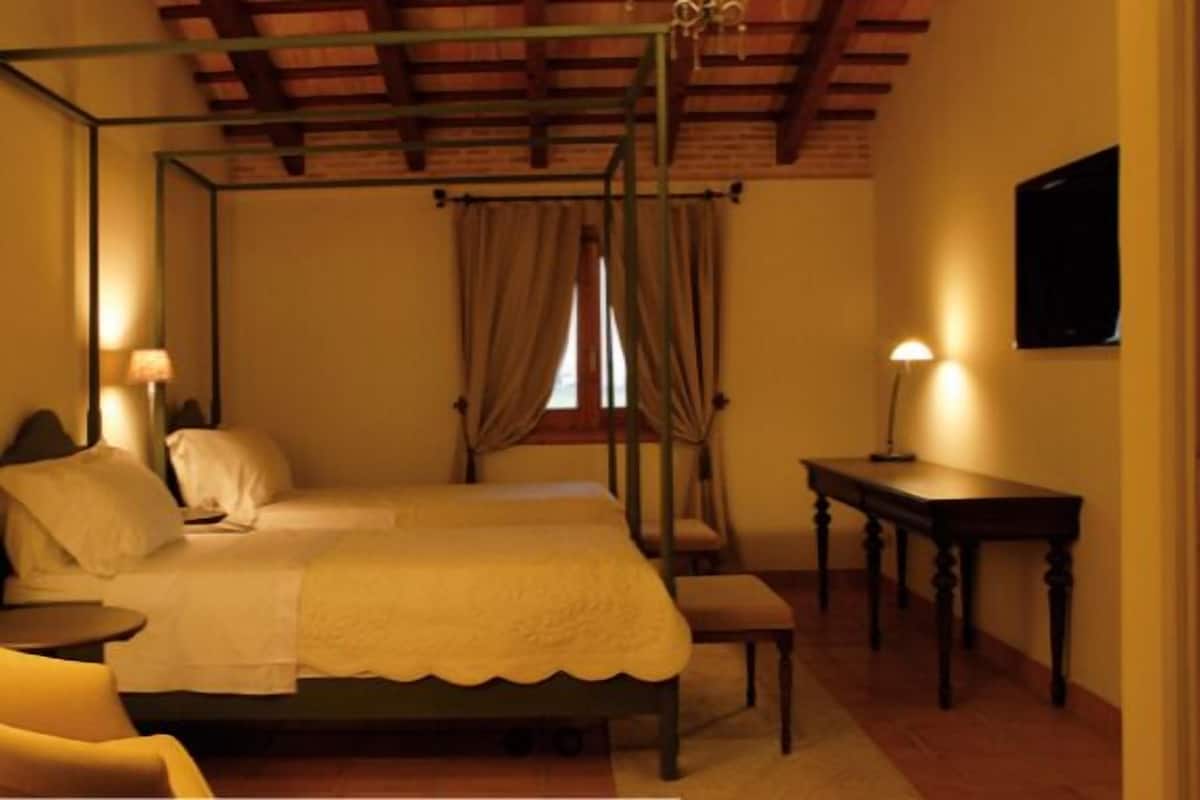 A four-poster bed with a canopy, a bedside table, a chair, and a desk in a warmly lit room with a wooden ceiling.