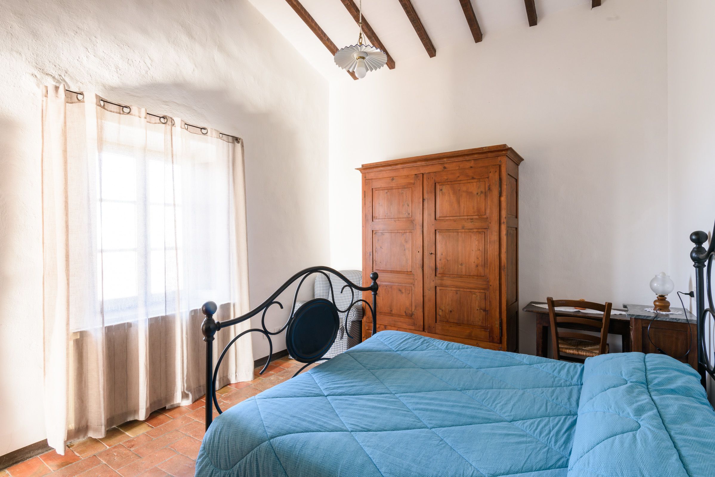 A bedroom with a bed, wooden wardrobe, a desk with a chair, and a window with curtains.