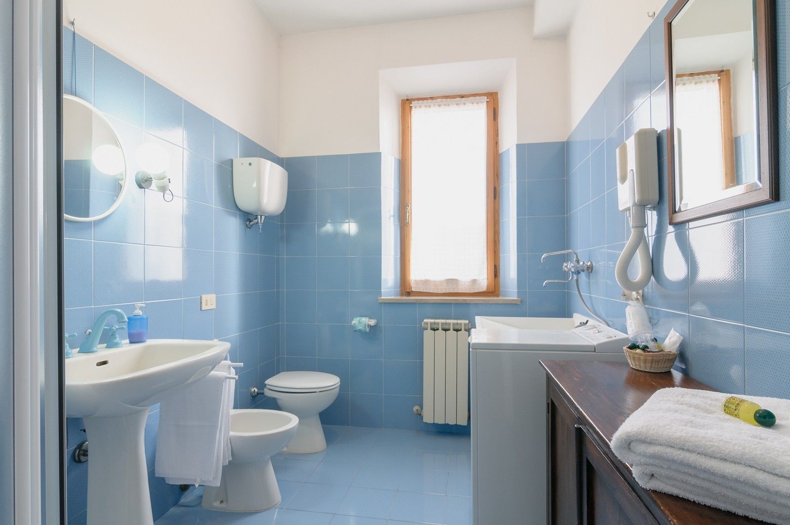 Ein Badezimmer mit blauen Fliesenwänden, einem weißen Waschbecken, einer Toilette, einem Spiegel und einem Fenster mit Vorhang.