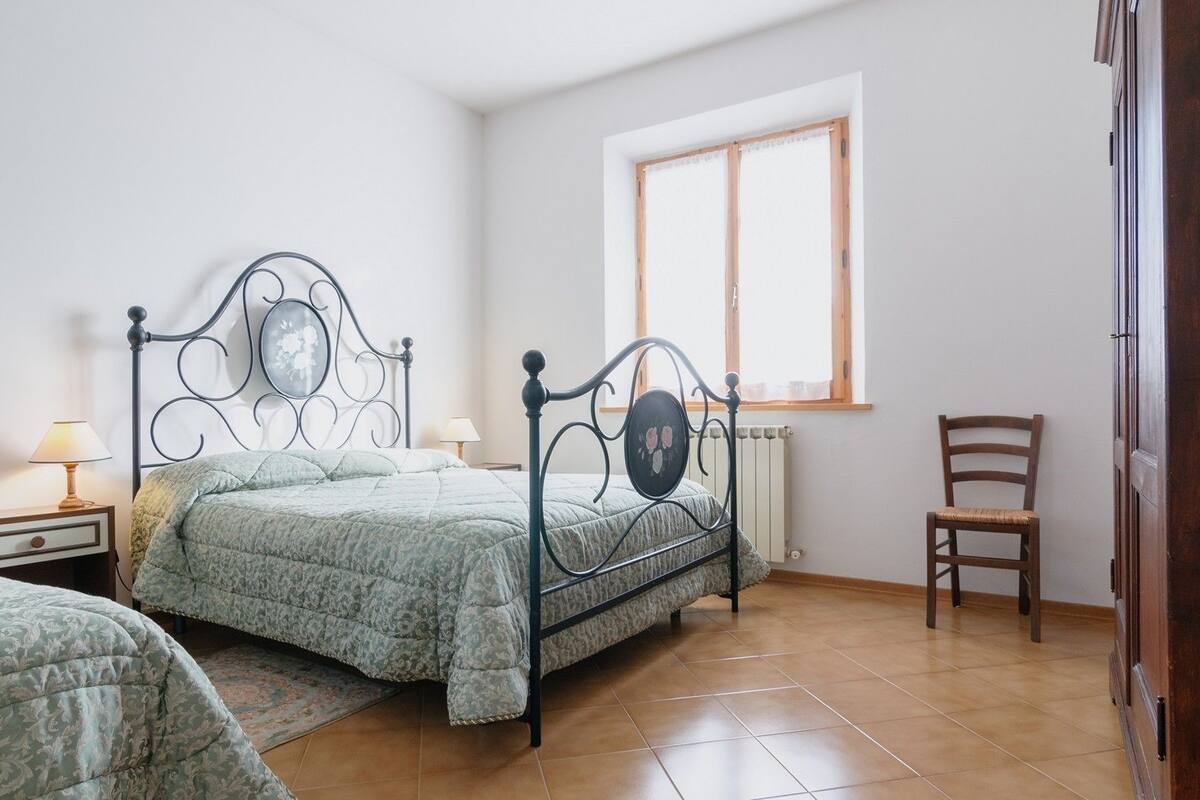 Un dormitorio con cama, dos mesitas de noche, una silla, una ventana y un radiador.