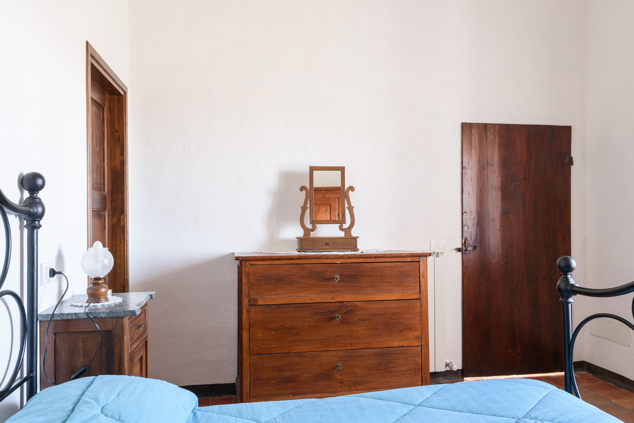 Een slaapkamer met een houten dressoir, een nachtkastje met een lamp, en een deur.