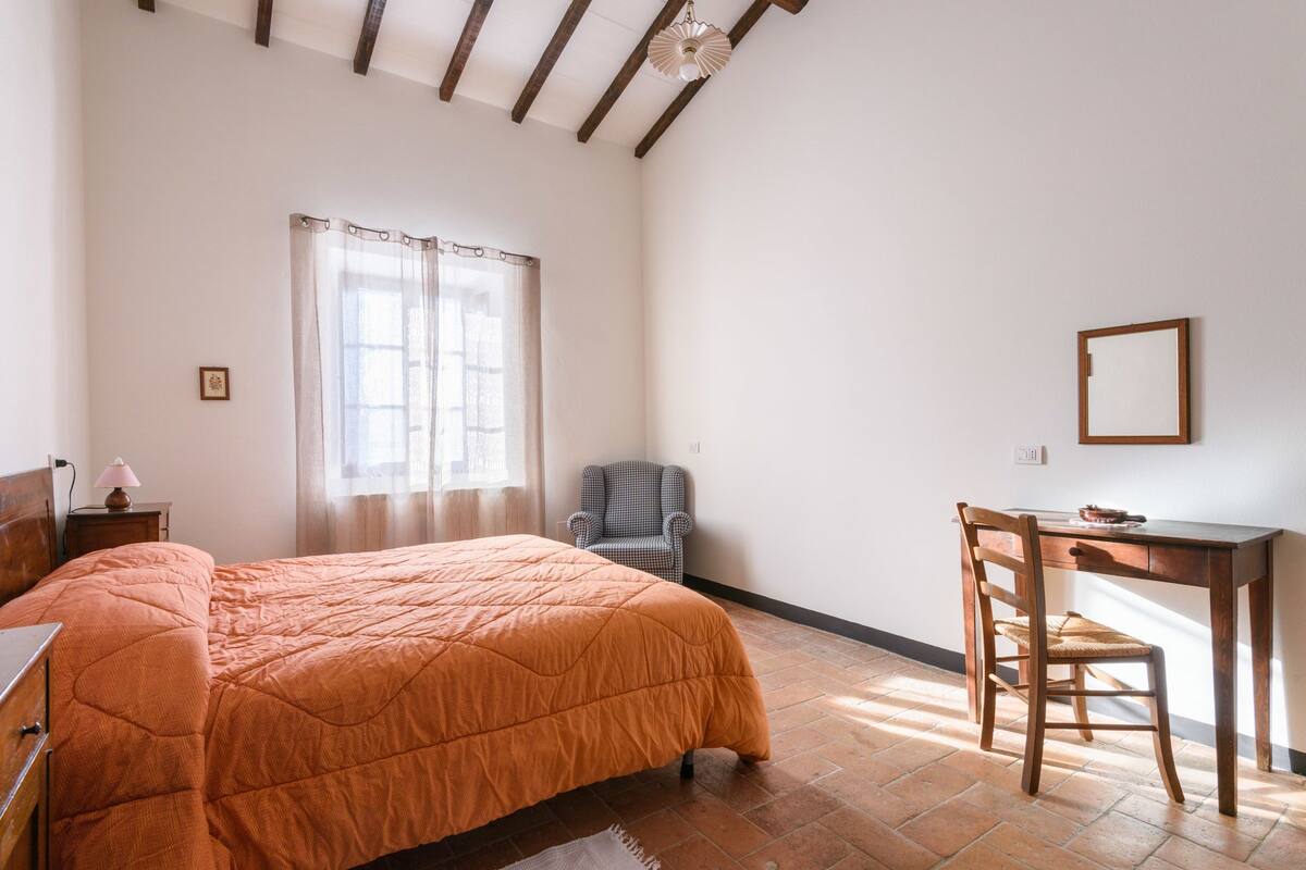 Un dormitorio con cama, una silla, un escritorio y una ventana con cortinas.