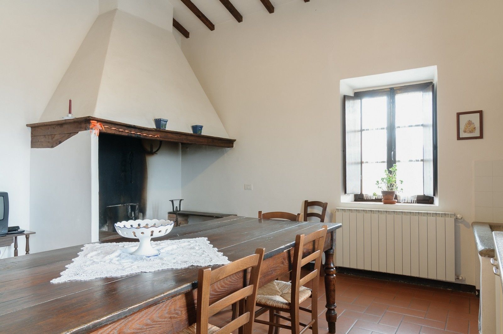 Een eethoek met een houten tafel, stoelen, een witte tafelloper van kant, een witte vaas en een raam met uitzicht op het omliggende landschap.
