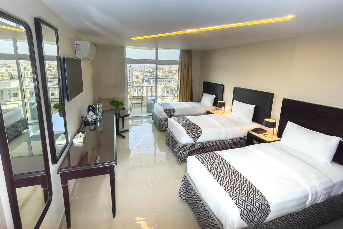 A hotel room with two beds, a desk, a chair, a TV, and a balcony with a city view.