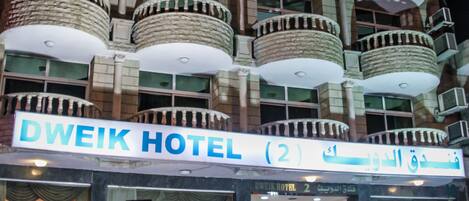 A multi-story hotel with balconies, a sign reading "DWEIK HOTEL (2)", and cars parked in front.