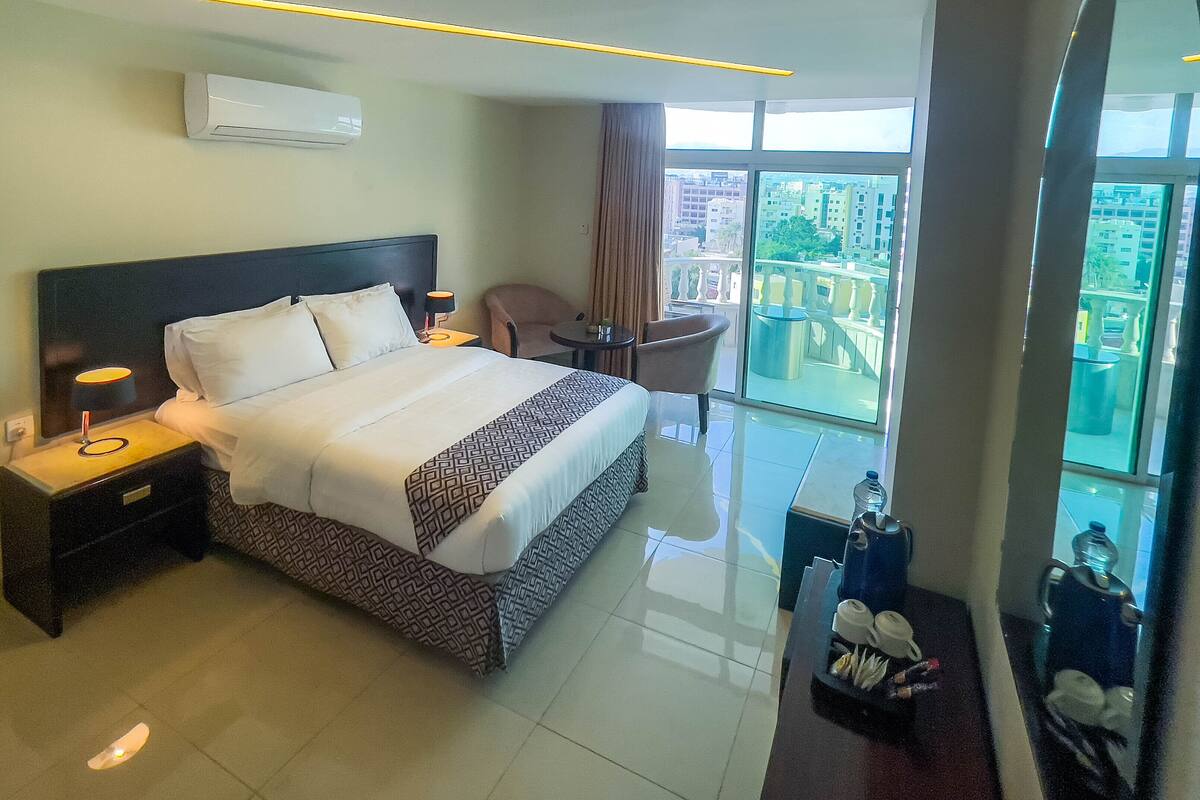 A hotel room with a large bed, a desk, a chair, and a view of the city through large windows.