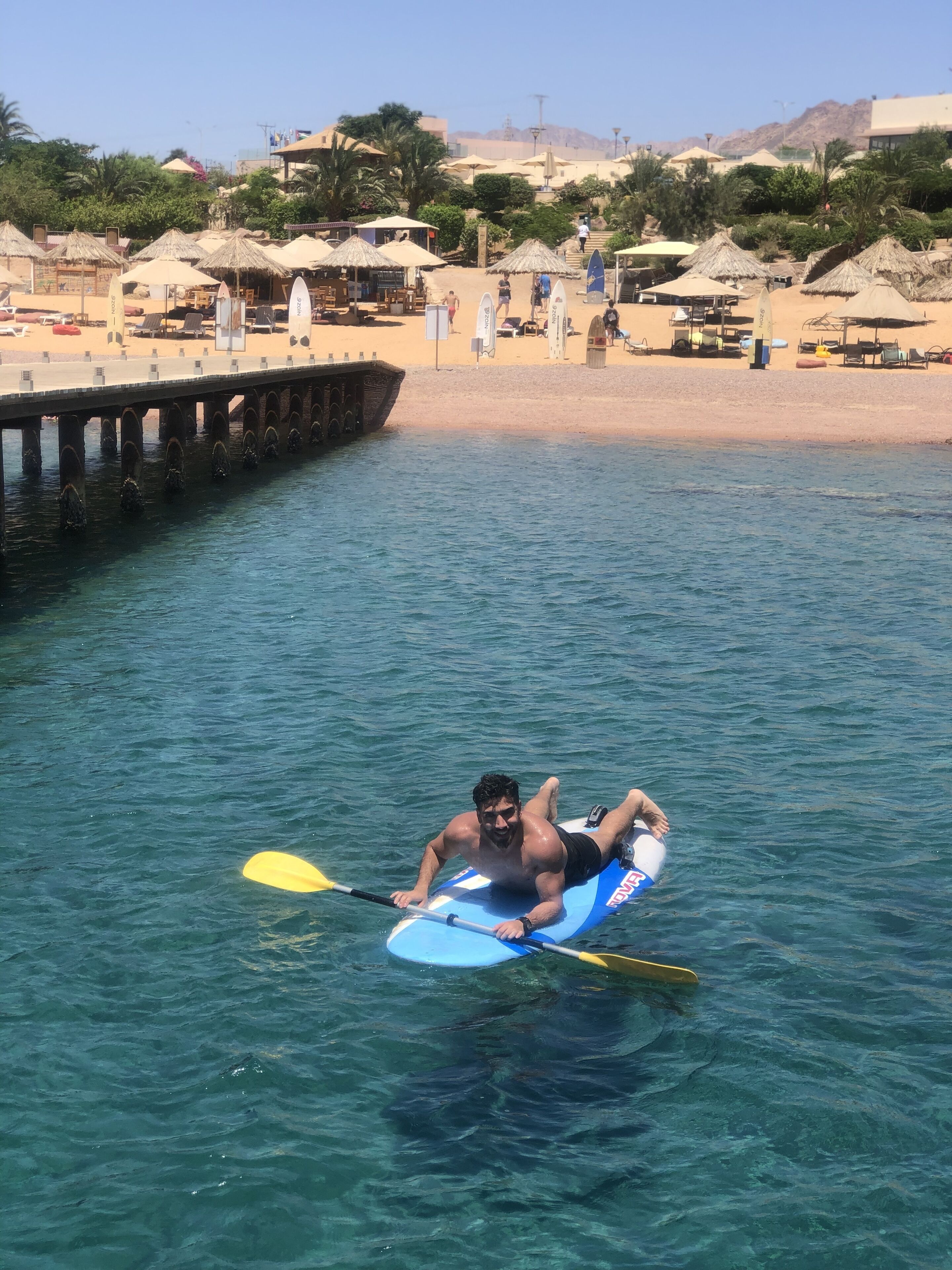 A man kayaking on a clear blue water body with a pier in the background.