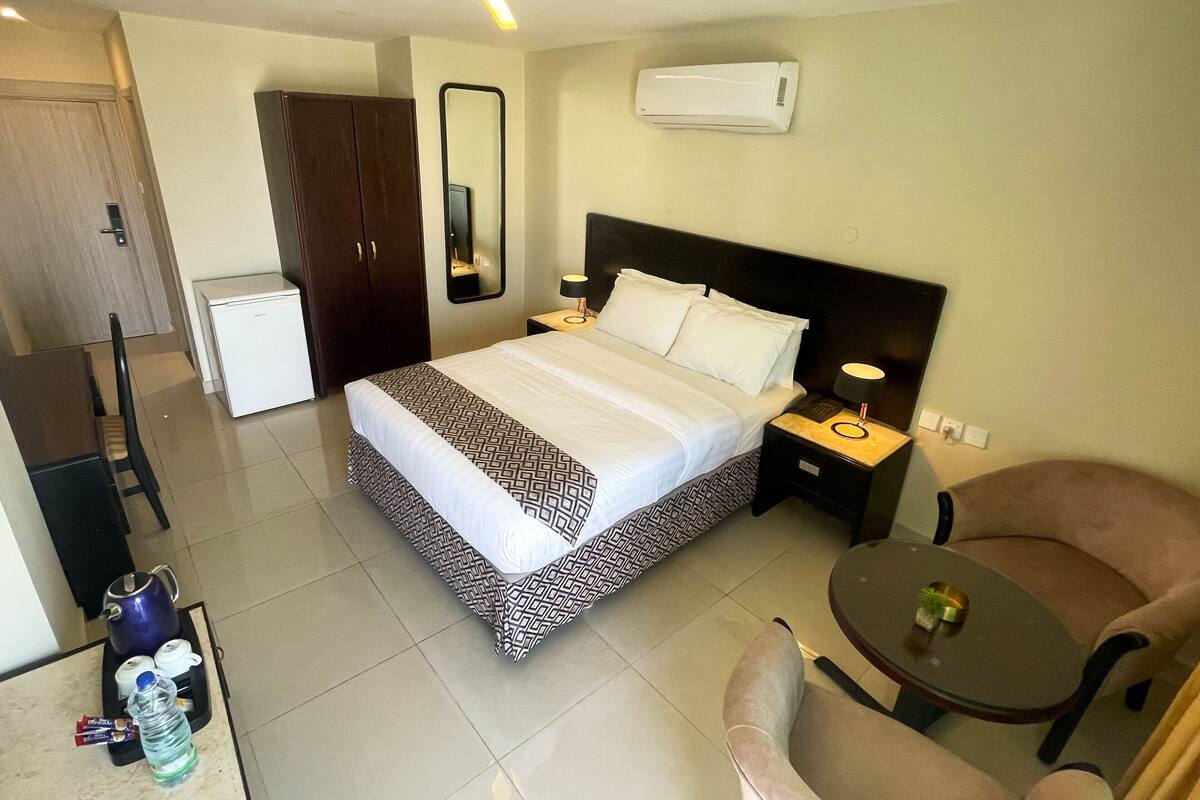 A hotel room with a bed, bedside table with lamp, chair, and a small table with a plant.