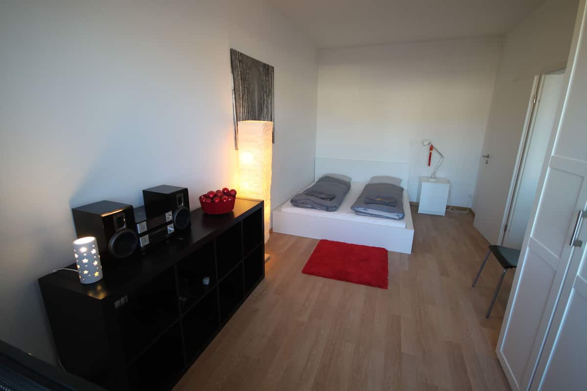 A bedroom with a bed, bedside table, lamp, and a black entertainment unit with speakers and a decorative item.
