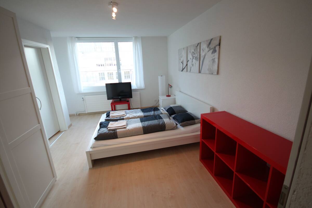 A modern bedroom with a bed, a TV, a window, and a red shelf unit.