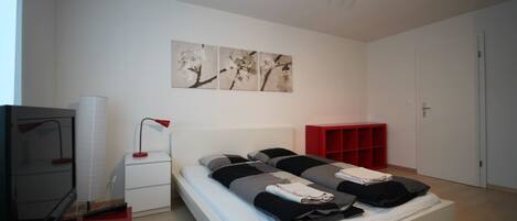 A modern bedroom with a bed, a TV, a desk, and a red shelving unit.