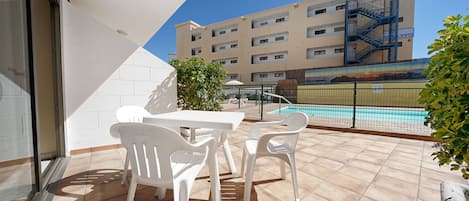 Un balcón con una mesa y sillas blancas, una piscina al fondo y un edificio con una escalera de incendios azul.