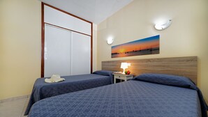 Een hotelkamer met twee bedden, een houten hoofdeinde, een nachtkastje met een lamp, een schilderij van een zonsondergang en een deur met houten frame.