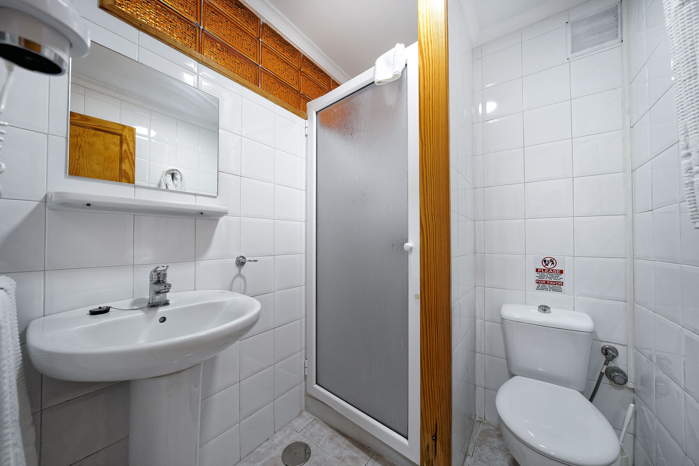 A bathroom with a white sink, mirror, and toilet, a shower enclosure, and a wooden panel above the mirror.