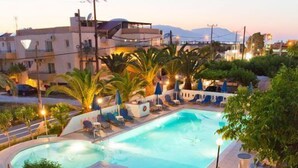 A swimming pool surrounded by lounge chairs and umbrellas, with a well-maintained lawn and a view of buildings in the background. - Anatoli Beach (Apokoronas)