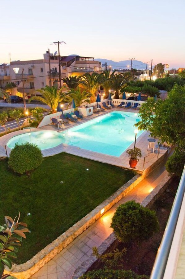 A swimming pool surrounded by lounge chairs and umbrellas, with a well-maintained lawn and a view of buildings in the background. - Anatoli Beach (Apokoronas)