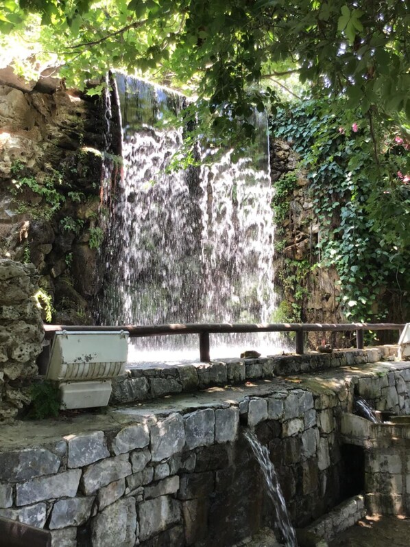 A waterfall cascading over a stone wall, surrounded by greenery and a wooden railing. - Anatoli Beach (Apokoronas)