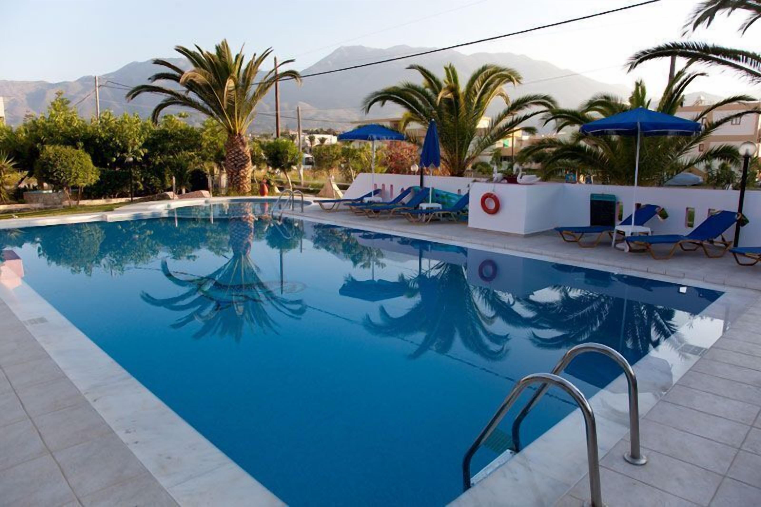 A swimming pool with clear blue water, surrounded by lounge chairs and umbrellas, with palm trees and mountains in the background.