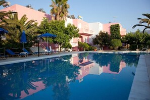 A swimming pool with lounge chairs and umbrellas, surrounded by palm trees and buildings with a pink facade. - Anatoli Beach (Apokoronas)