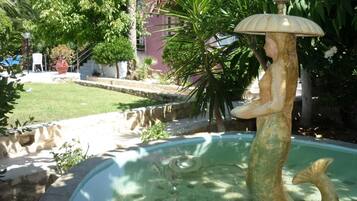 Una fuente con una estatua de sirena, rodeada de follaje y un edificio al fondo.