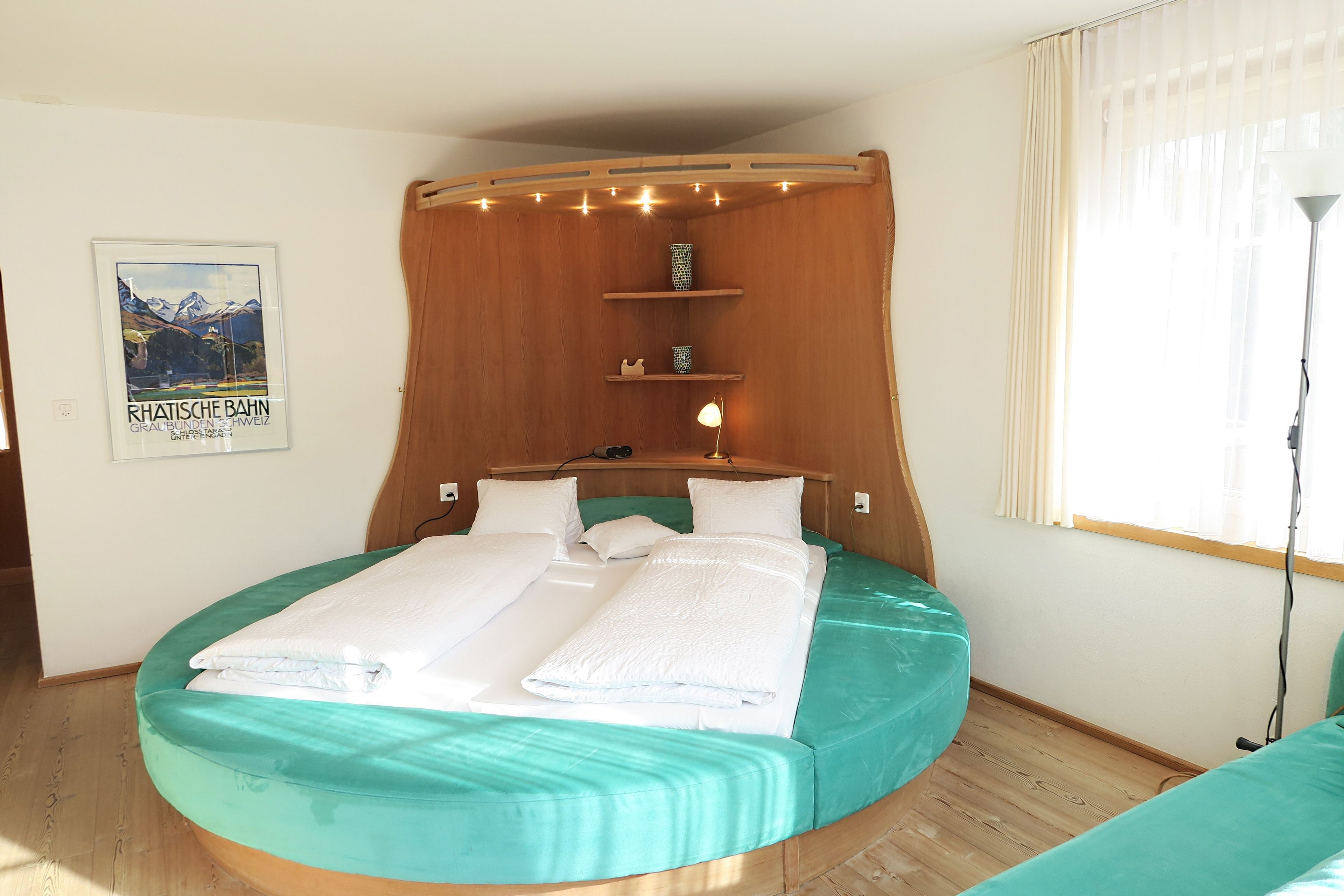 Ein rundes Bett mit einem Holz-Kopfteil, weißen Bettwäsche und einer türkisblauen Tagesdecke.