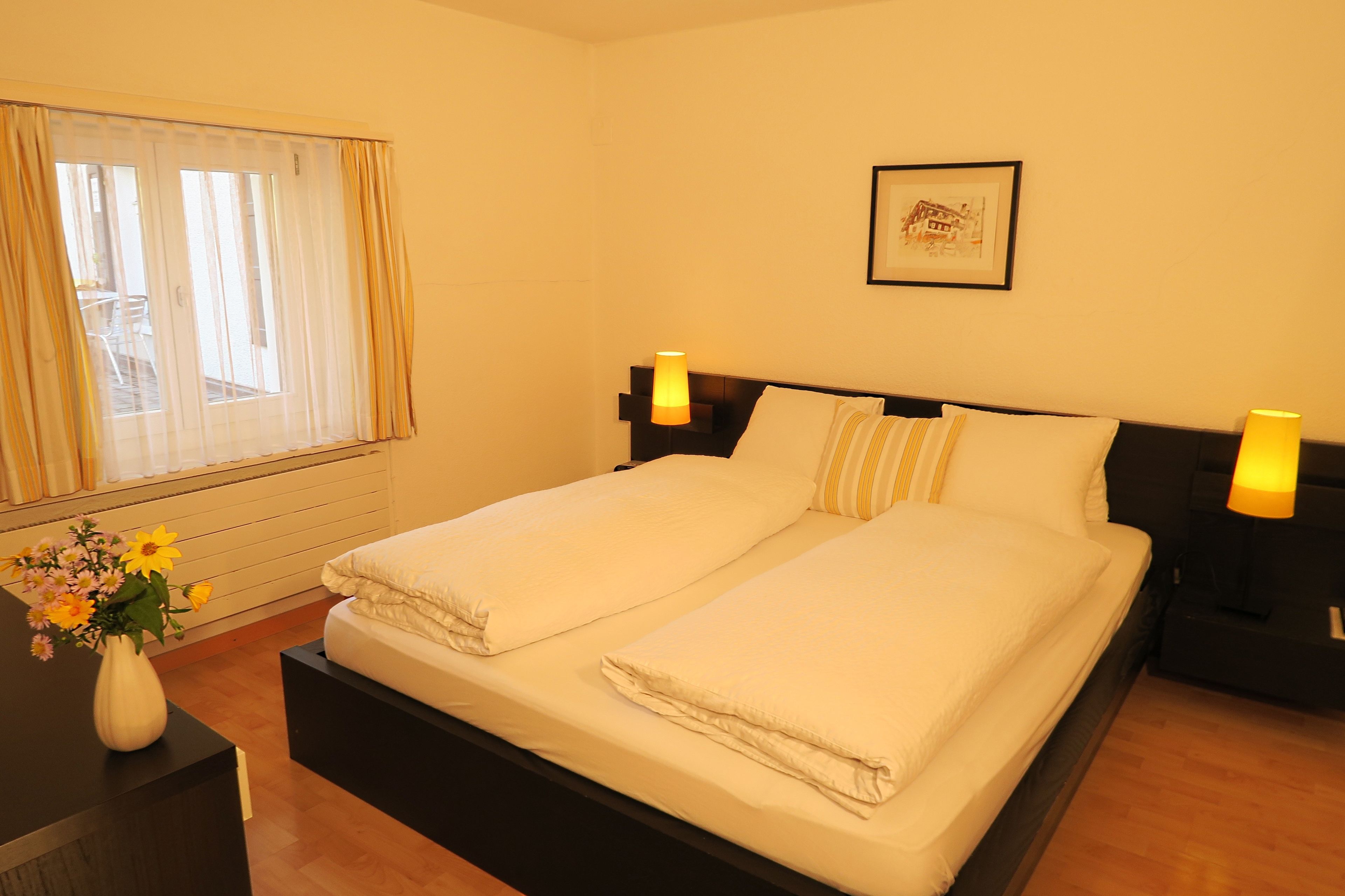 A double bed with white linens, two bedside lamps, a framed picture on the wall, and a window with curtains.