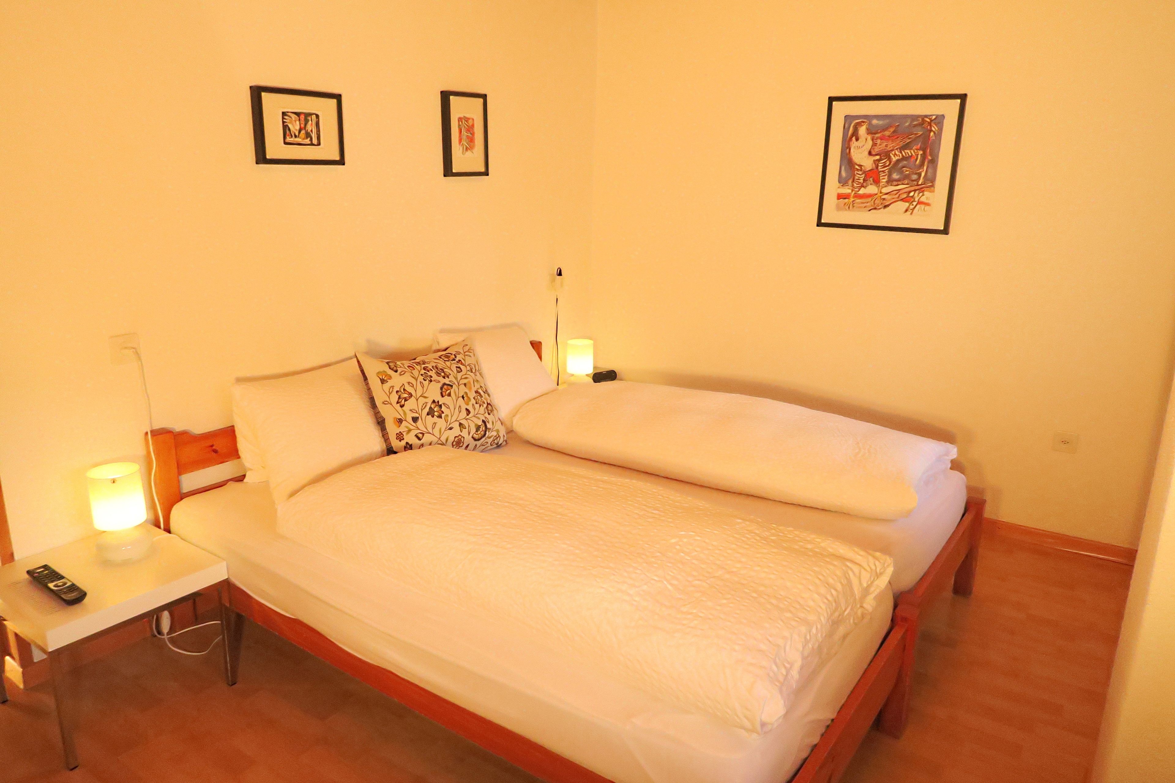 A neatly made bed with white linens, a bedside table with a lamp, and three framed pictures on the wall.