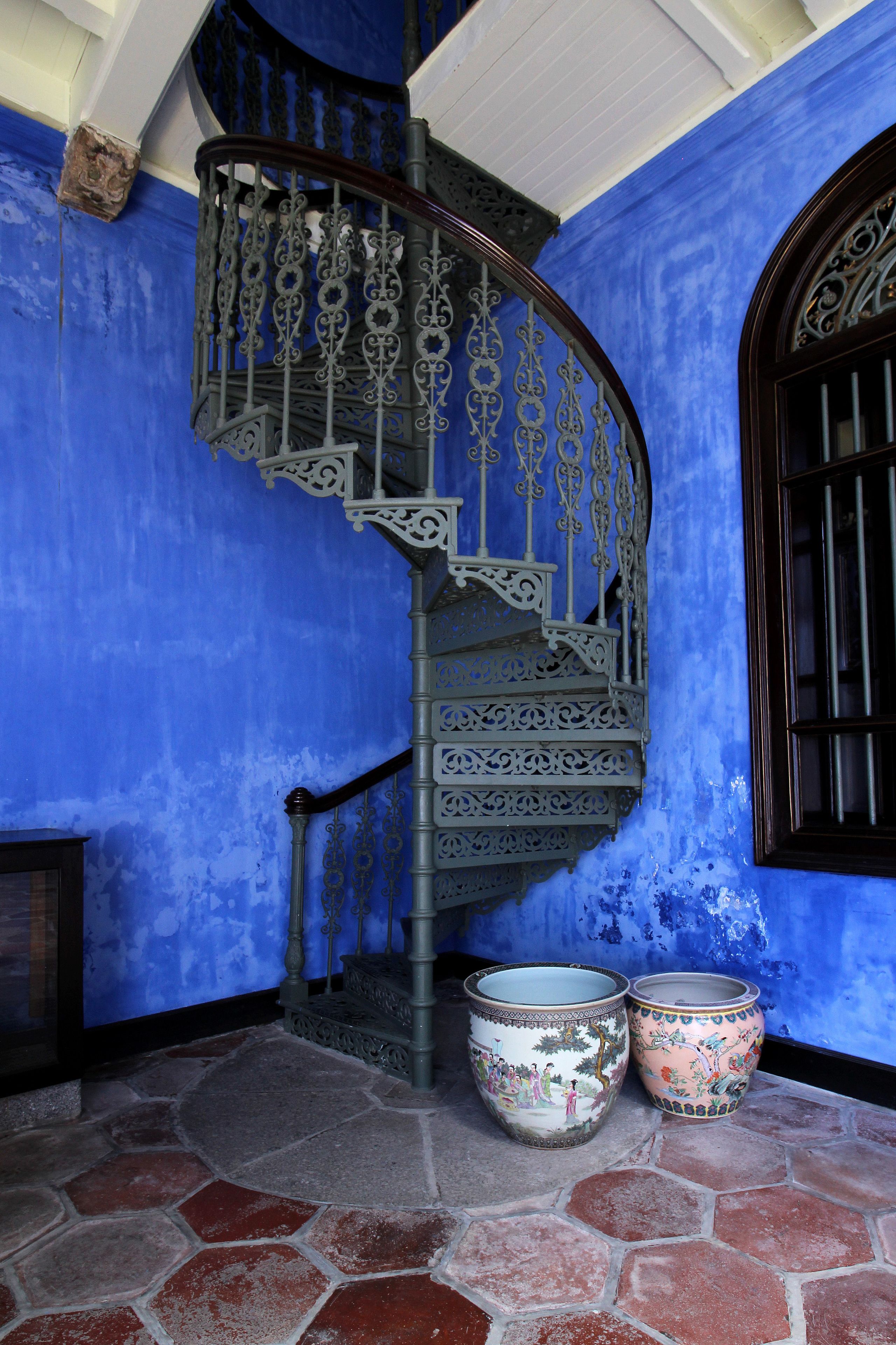 A spiral staircase with ornate metal railings, two ceramic pots with floral designs, and a blue wall with a window with bars.