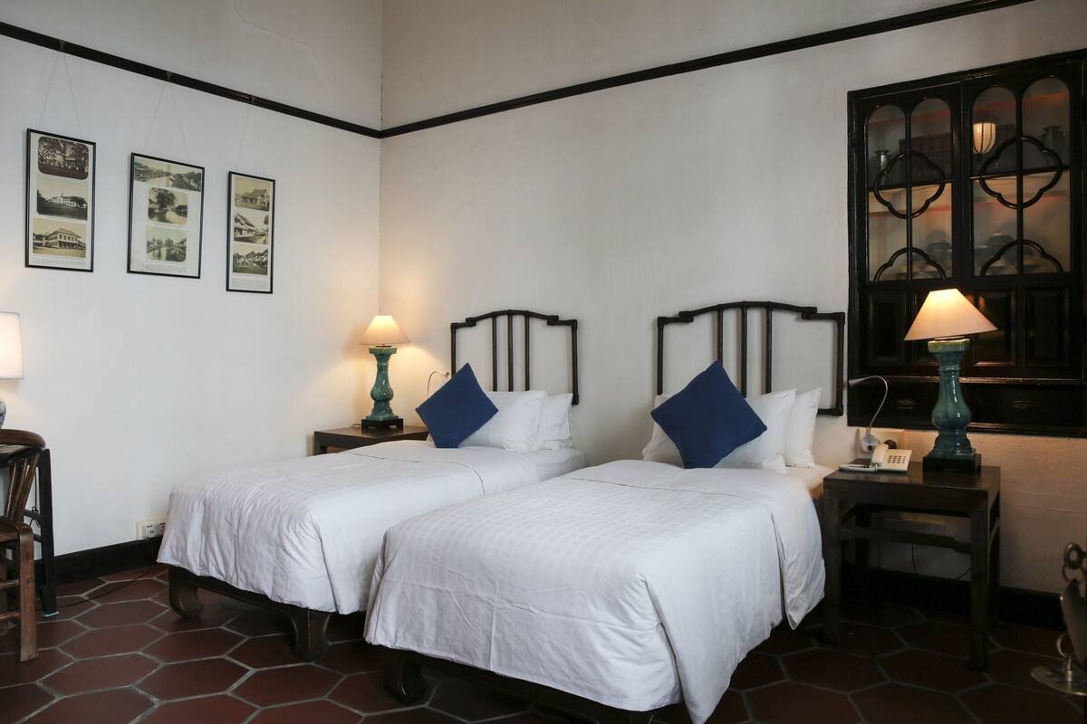 A room with two beds, a nightstand with a lamp, a window with a decorative frame, and three framed pictures on the wall.