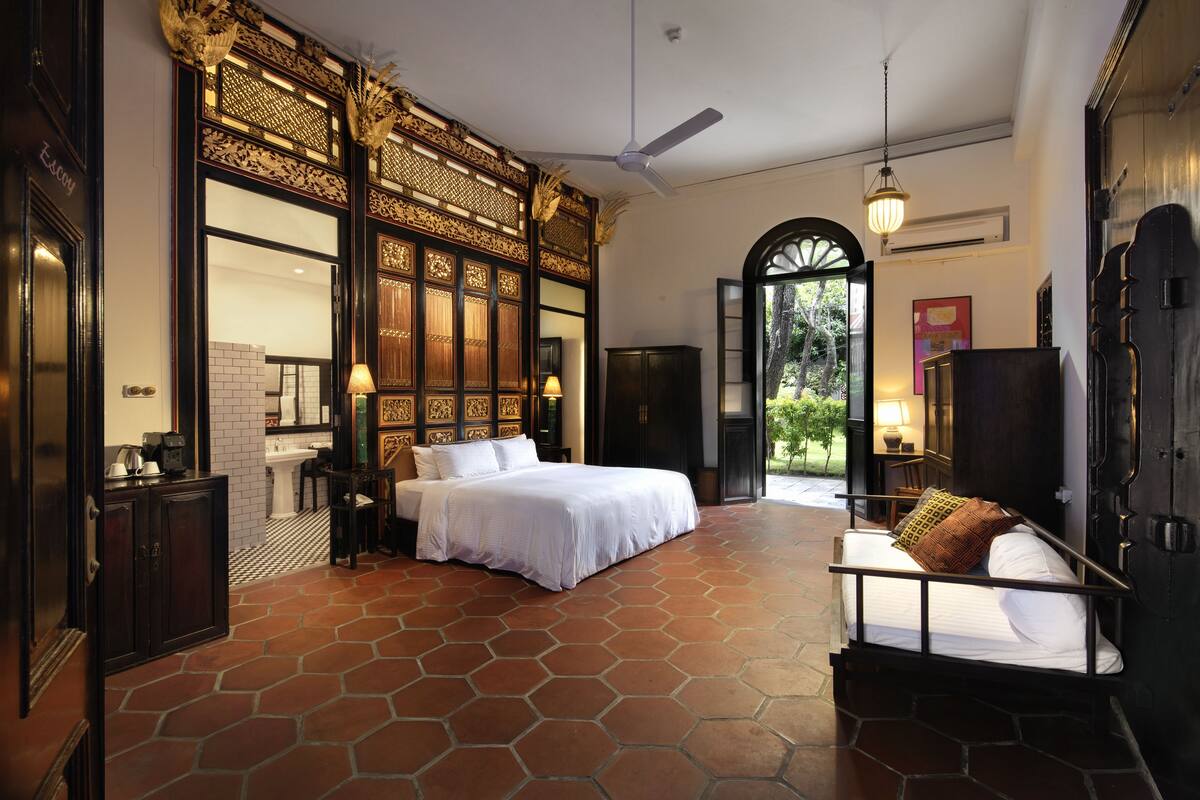 A spacious bedroom with a large bed, a sofa, and a view of the garden through an open door.