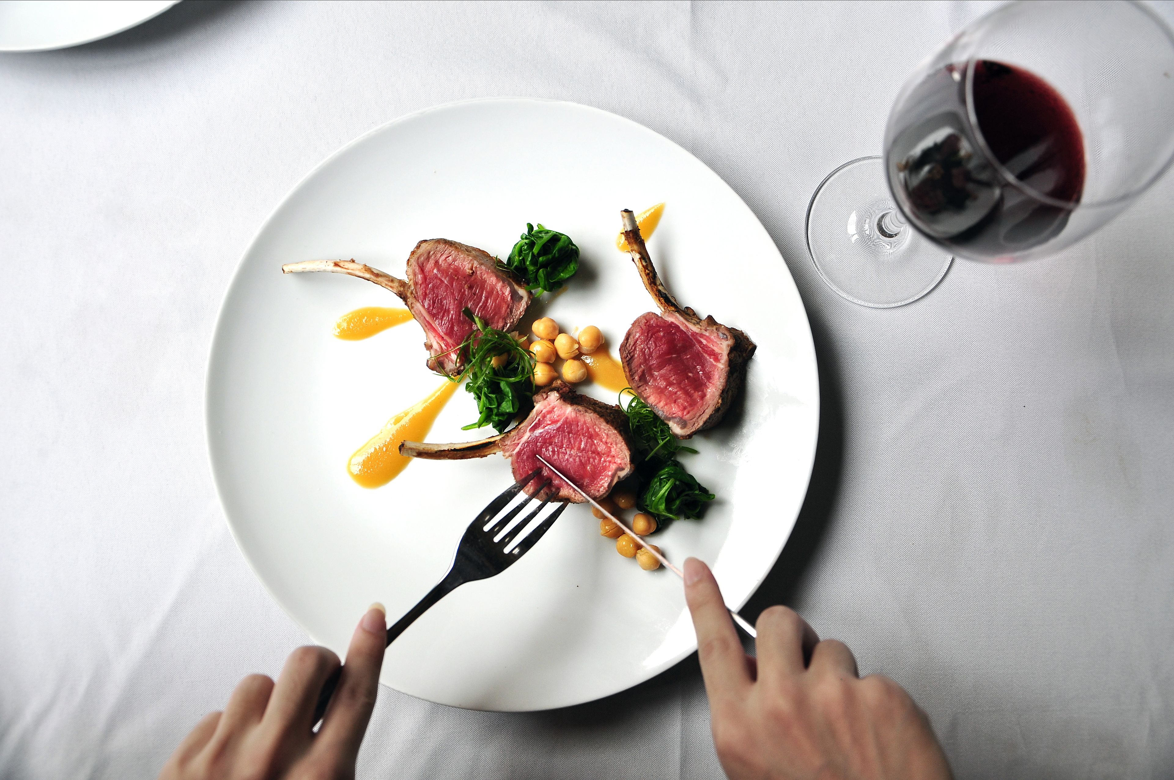 A white plate with a steak, greens, and a sauce, accompanied by a fork and a glass of red wine.