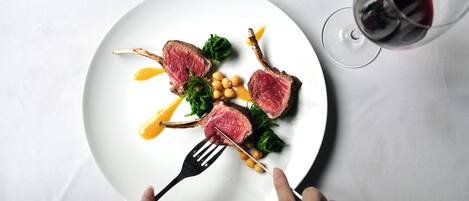 A white plate with a steak, greens, and a sauce, accompanied by a fork and a glass of red wine.