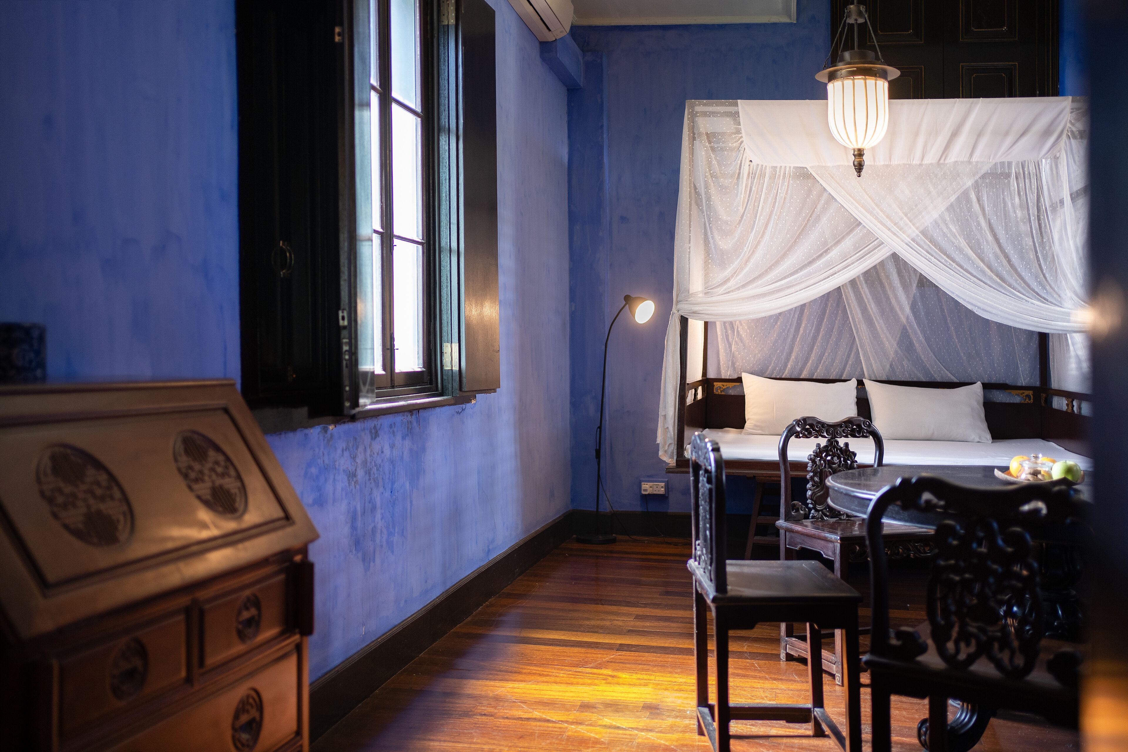 A bedroom with a bed under a white canopy, a wooden dresser, a desk with a chair, and a window with a view.