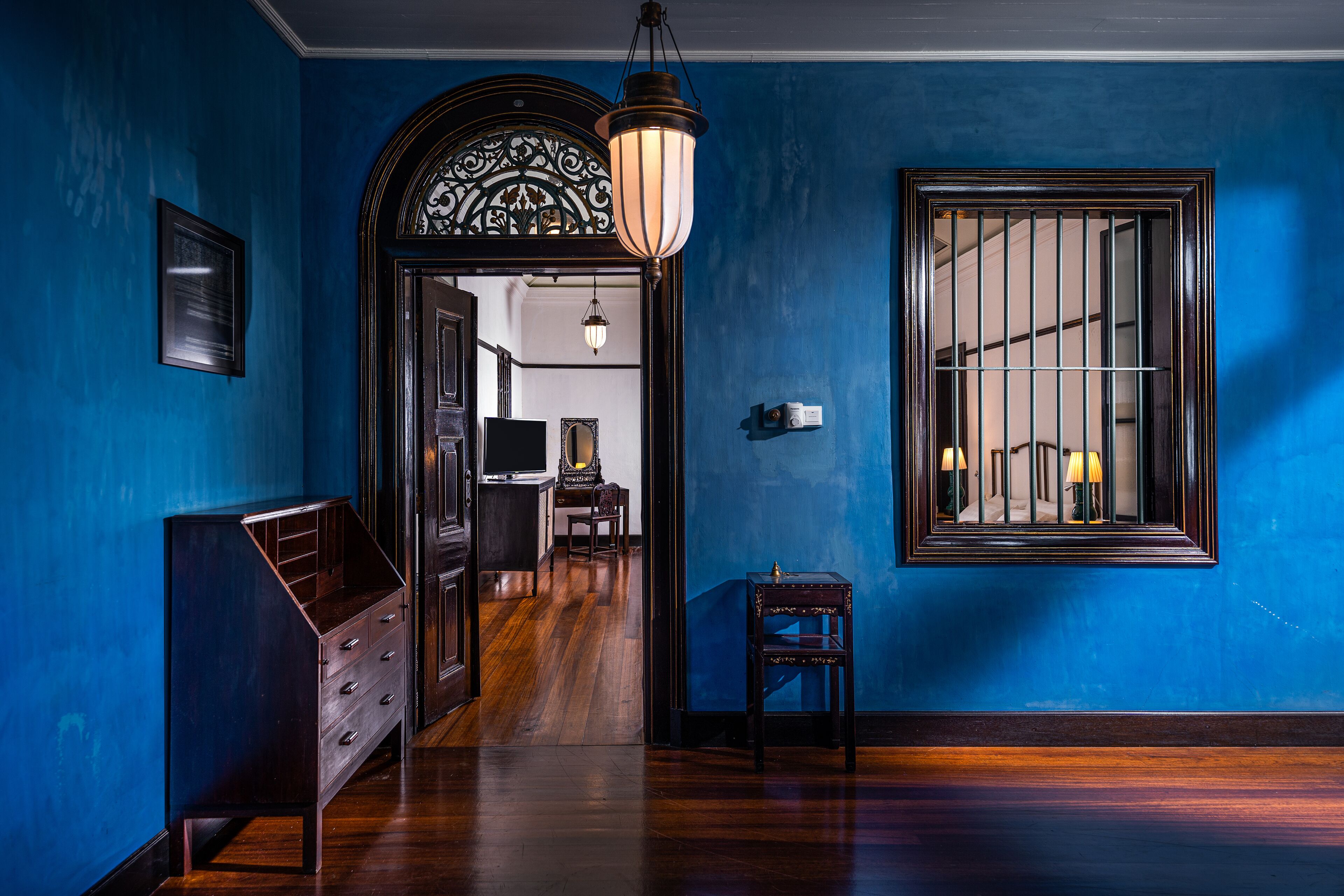 A hallway with a blue wall, wooden floor, a dark wooden dresser, a small table, and a window with bars.