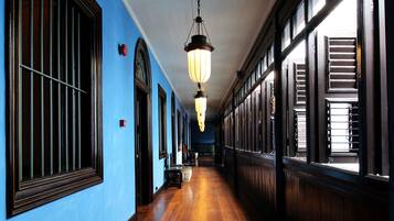 Un long couloir au sol en bois et aux murs bleus, agrémenté de grandes fenêtres avec volets et d’un luminaire suspendu.