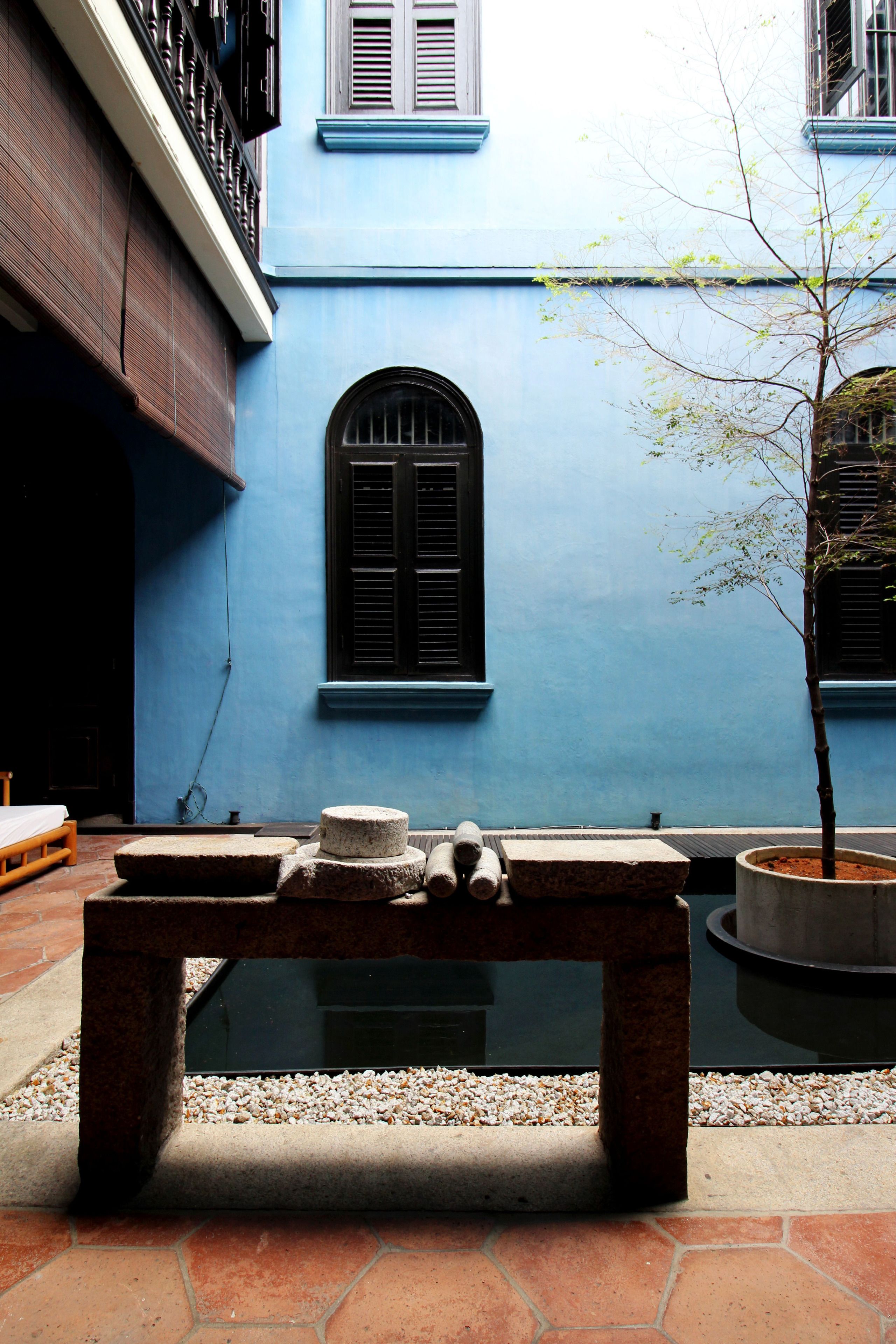 A courtyard with a bench, a small tree, and a building with blue walls and a window.