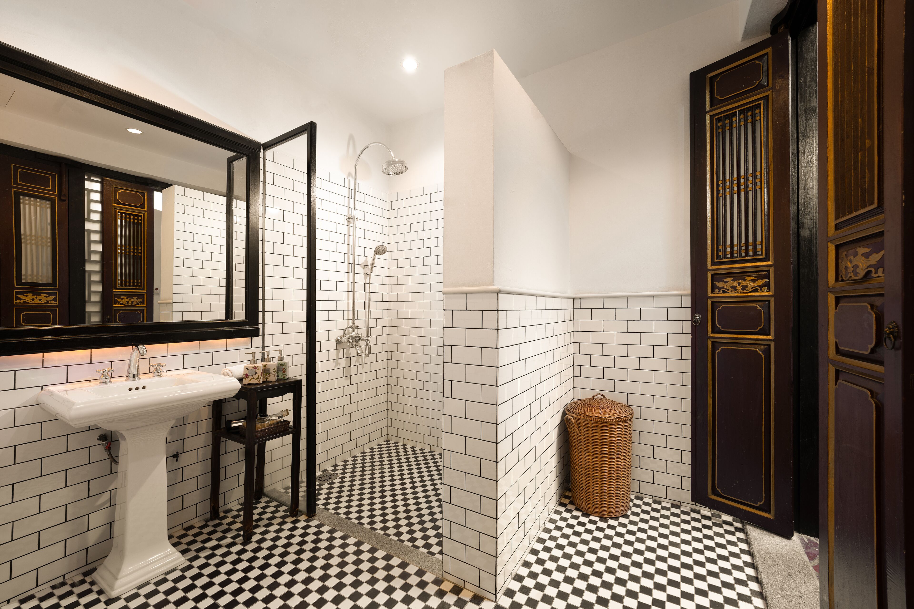 Un baño con un lavamanos blanco de pedestal, una ducha en esquina con piso a cuadros blanco y negro, y una puerta de madera oscura con detalles dorados.