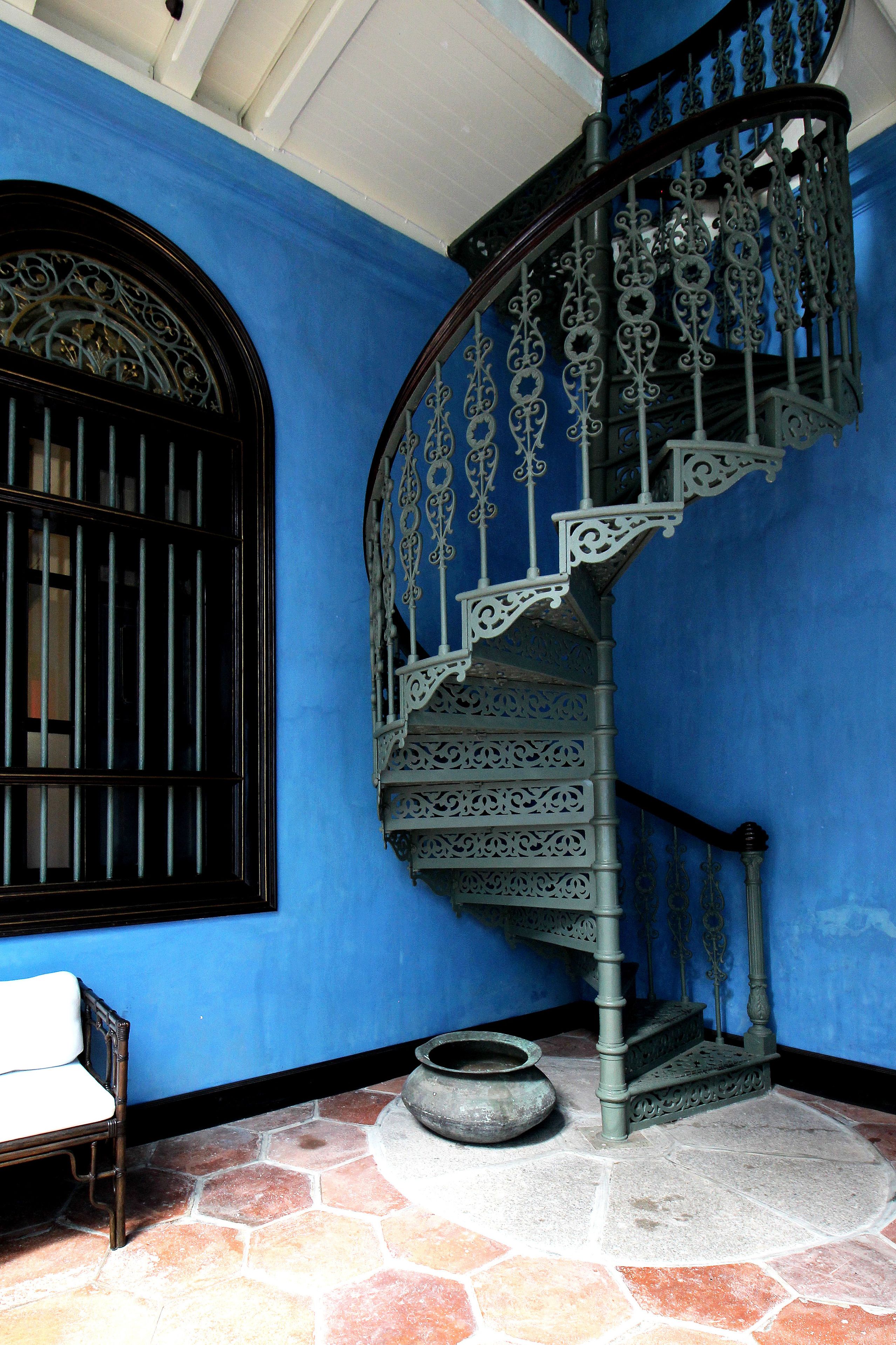 A spiral staircase with ornate metal railings, a window with bars, and a stone floor.