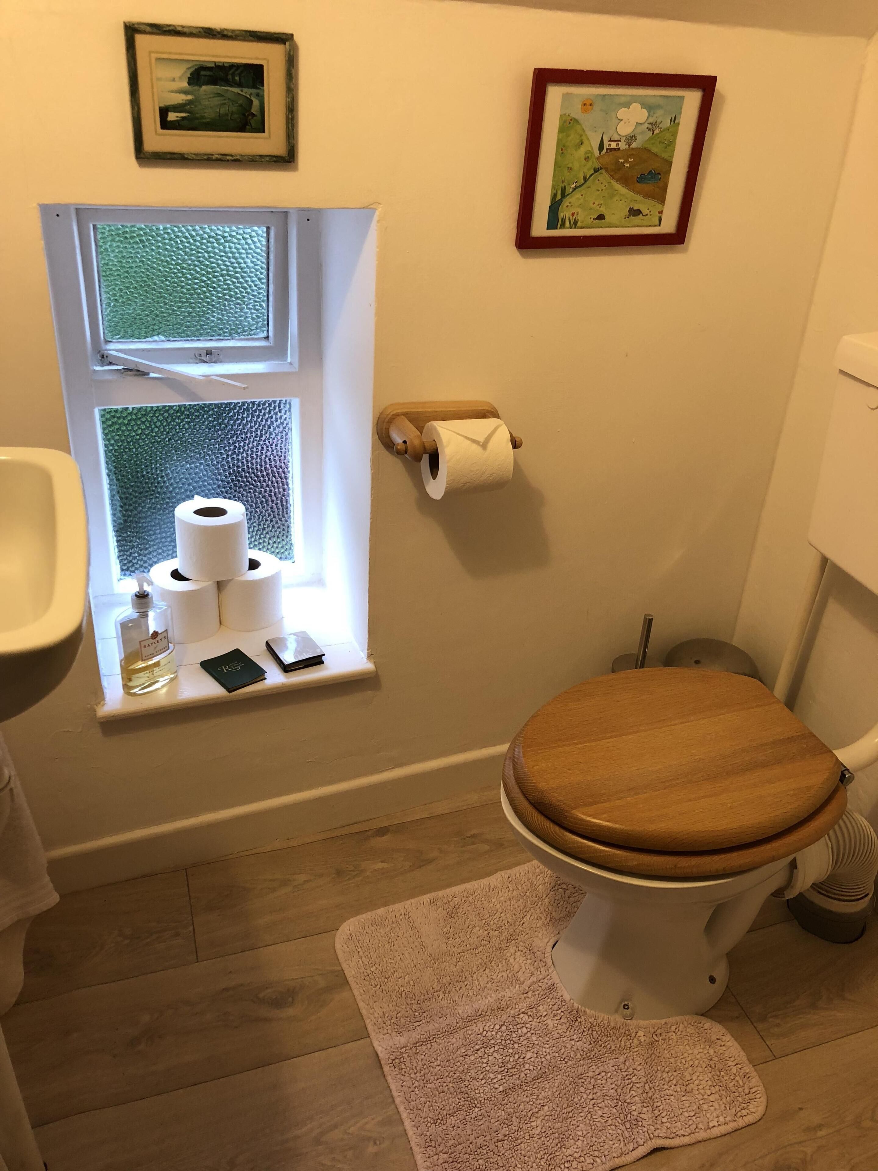 Une salle de bain avec des toilettes, une fenêtre avec du papier toilette, une petite étagère avec une bouteille et un téléphone, et deux tableaux encadrés accrochés au mur.