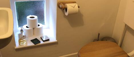 Une salle de bain avec des toilettes, une fenêtre avec du papier toilette, une petite étagère avec une bouteille et un téléphone, et deux tableaux encadrés accrochés au mur.