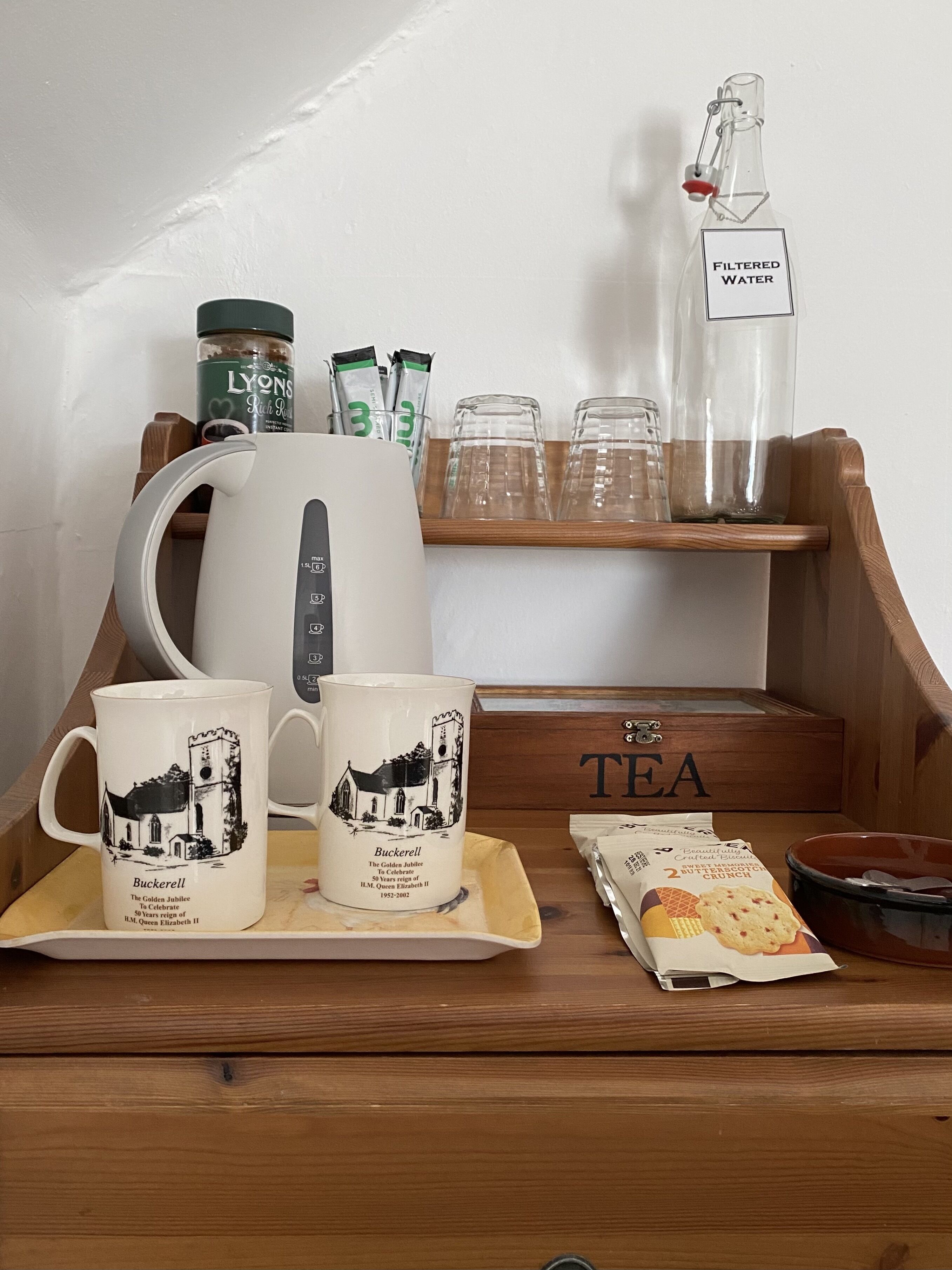 Una bandeja de madera con un hervidor de agua, dos tazas, una botella de vidrio con agua filtrada, bolsitas de té y una caja de galletas.